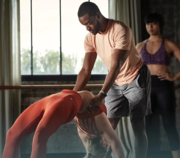 Fitness instructor assists a woman in a backbend stretch during a studio class while another participant watches.