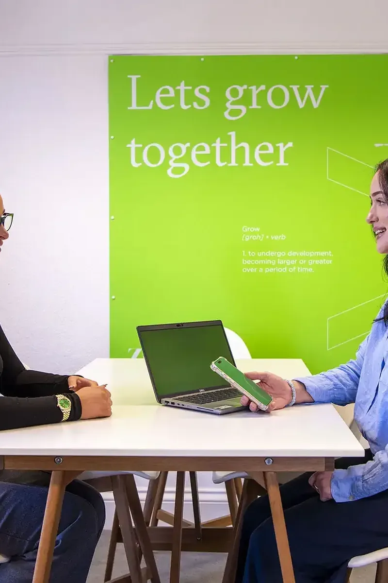Two women sat opposite each other on a white desk, one holding a phone