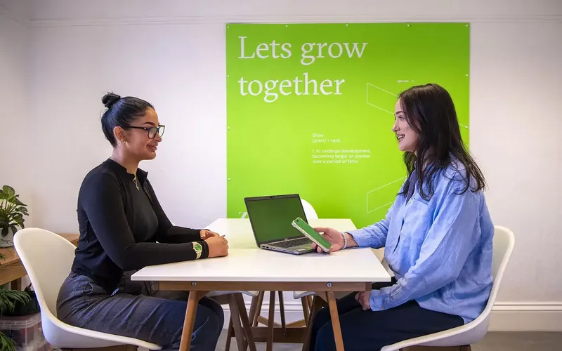 Two women sat opposite each other on a white desk, one holding a phone