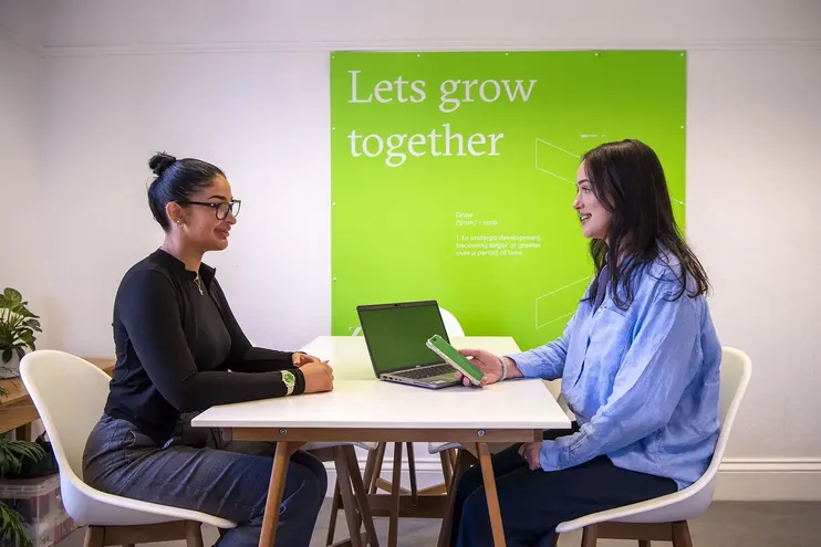 Two women sat opposite each other on a white desk, one holding a phone