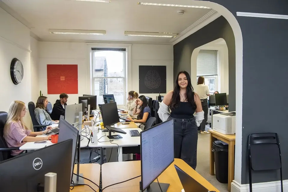 Office room with computers and people working on the computers, one woman stood behind wooden desk looking forward smiling